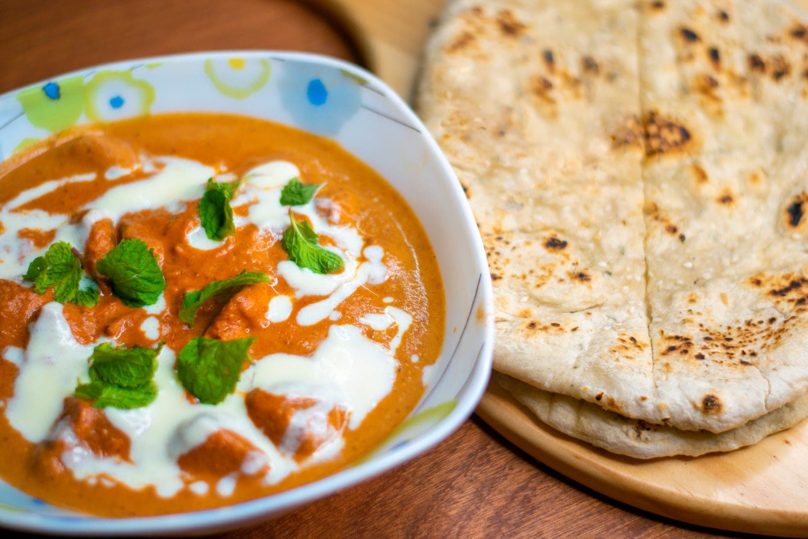 Chicken tikka masala in a creamy orange gravy with swirls of cream and fresh mint, beside naan on a wooden board