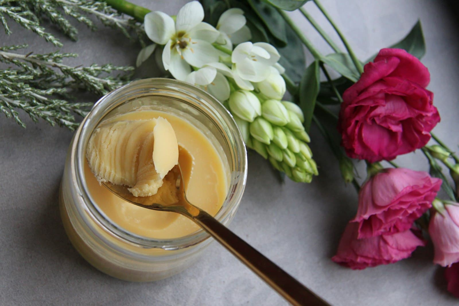 A glass jar of set ghee with a spoonful lifted out, beside white and pink flowers