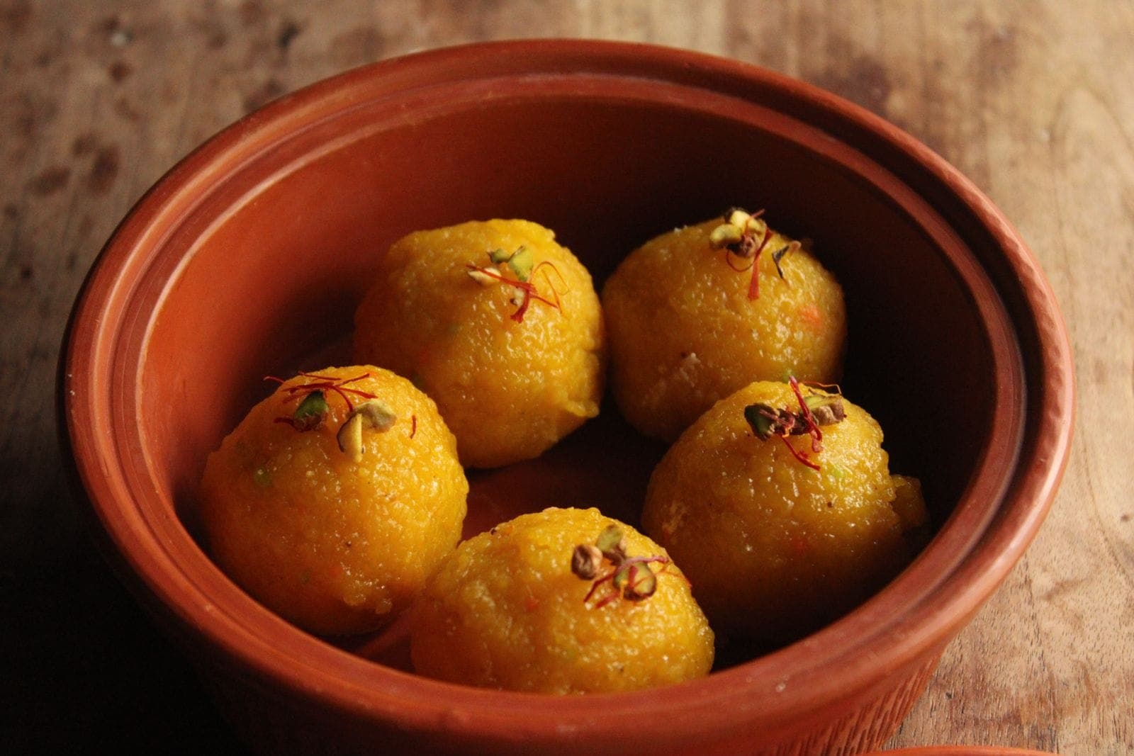 Five round yellow ladoos topped with saffron and pistachio in a clay bowl