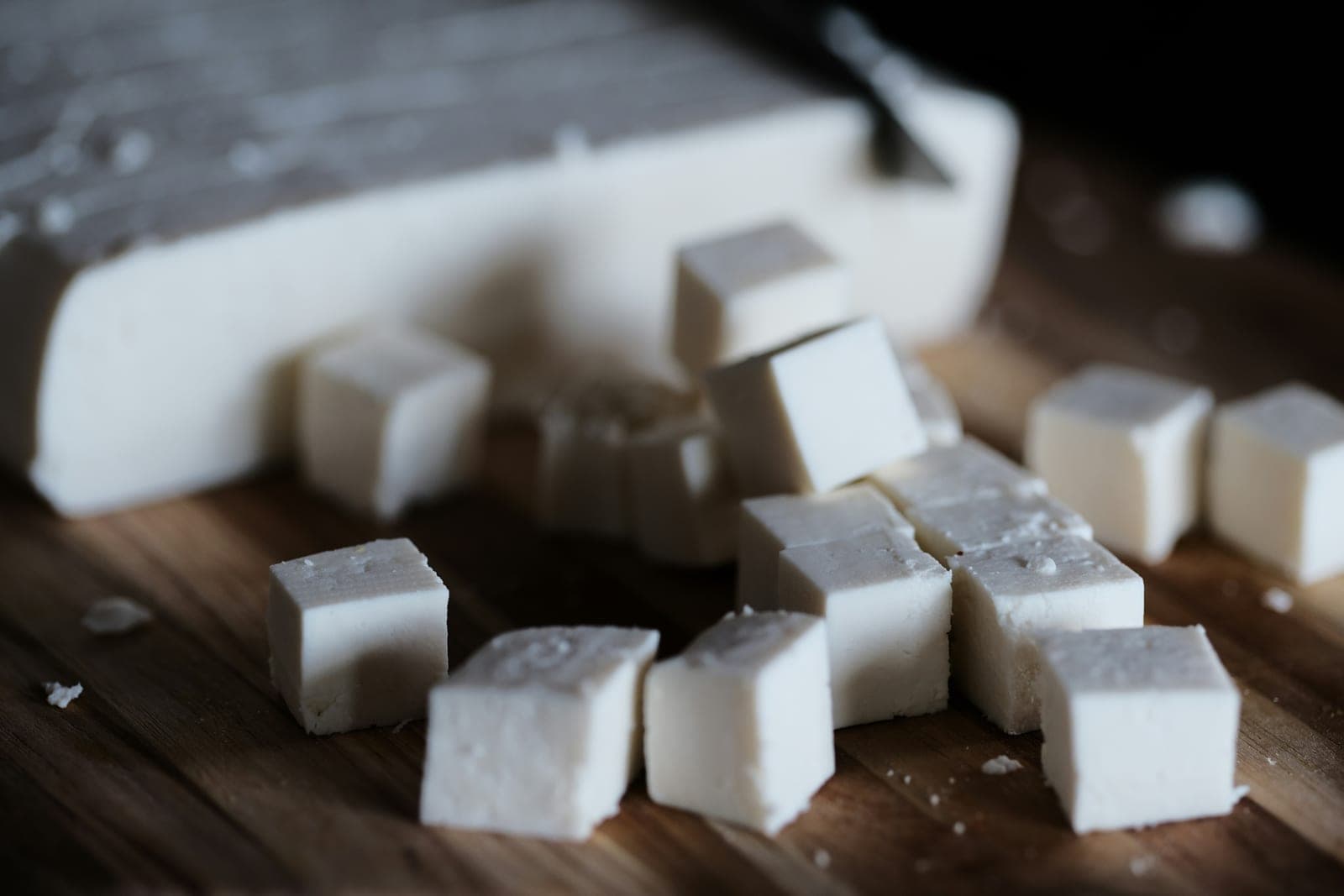 A block of white paneer on a wooden board, with part of it cut into cubes