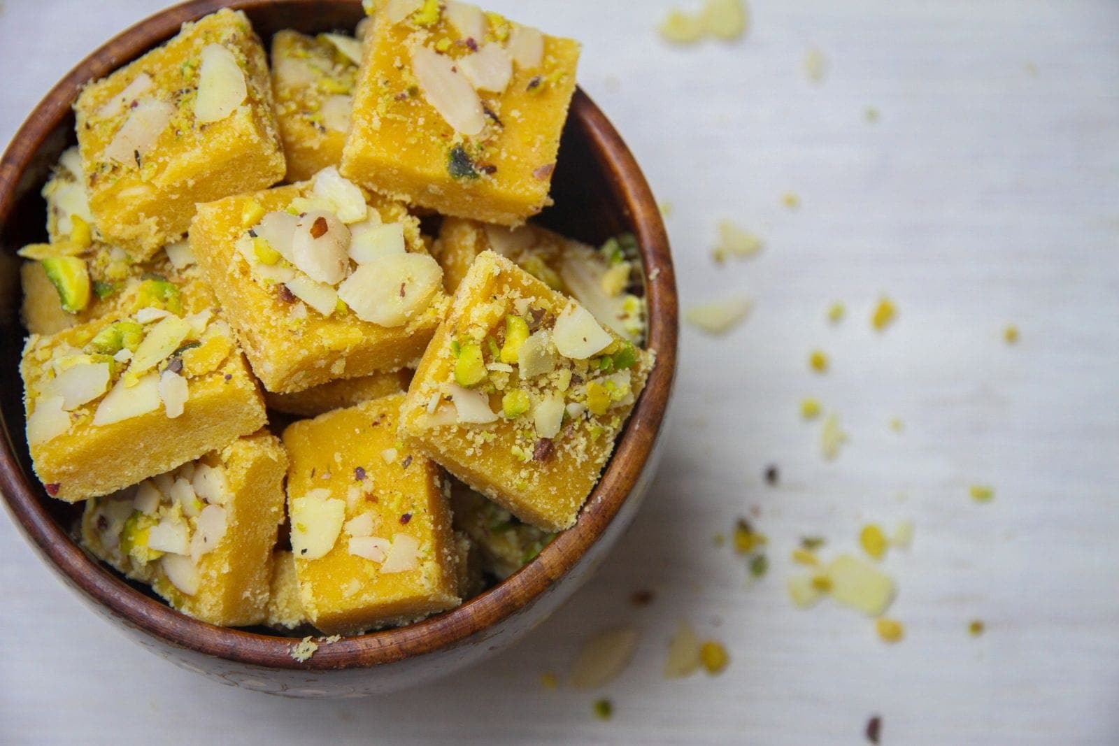 Squares of besan barfi topped with sliced almonds and pistachios in a wooden bowl